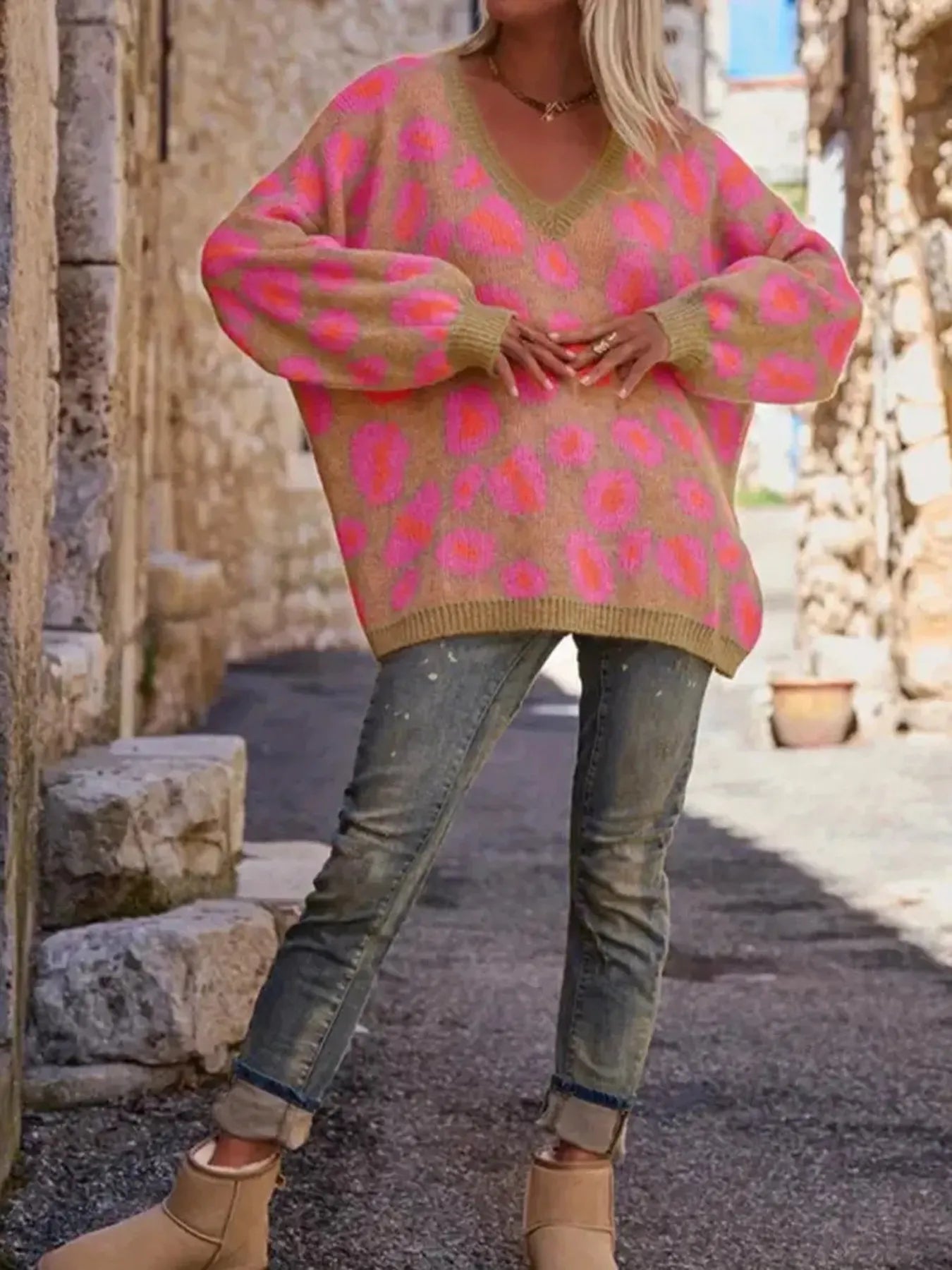Woman wearing a Leopard Oversized V-Neck Sweater with pink leopard print, paired with jeans and beige boots, in a rustic outdoor setting.