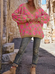 Woman wearing a Leopard Oversized V-Neck Sweater with pink leopard print, paired with jeans and beige boots, in a rustic outdoor setting.