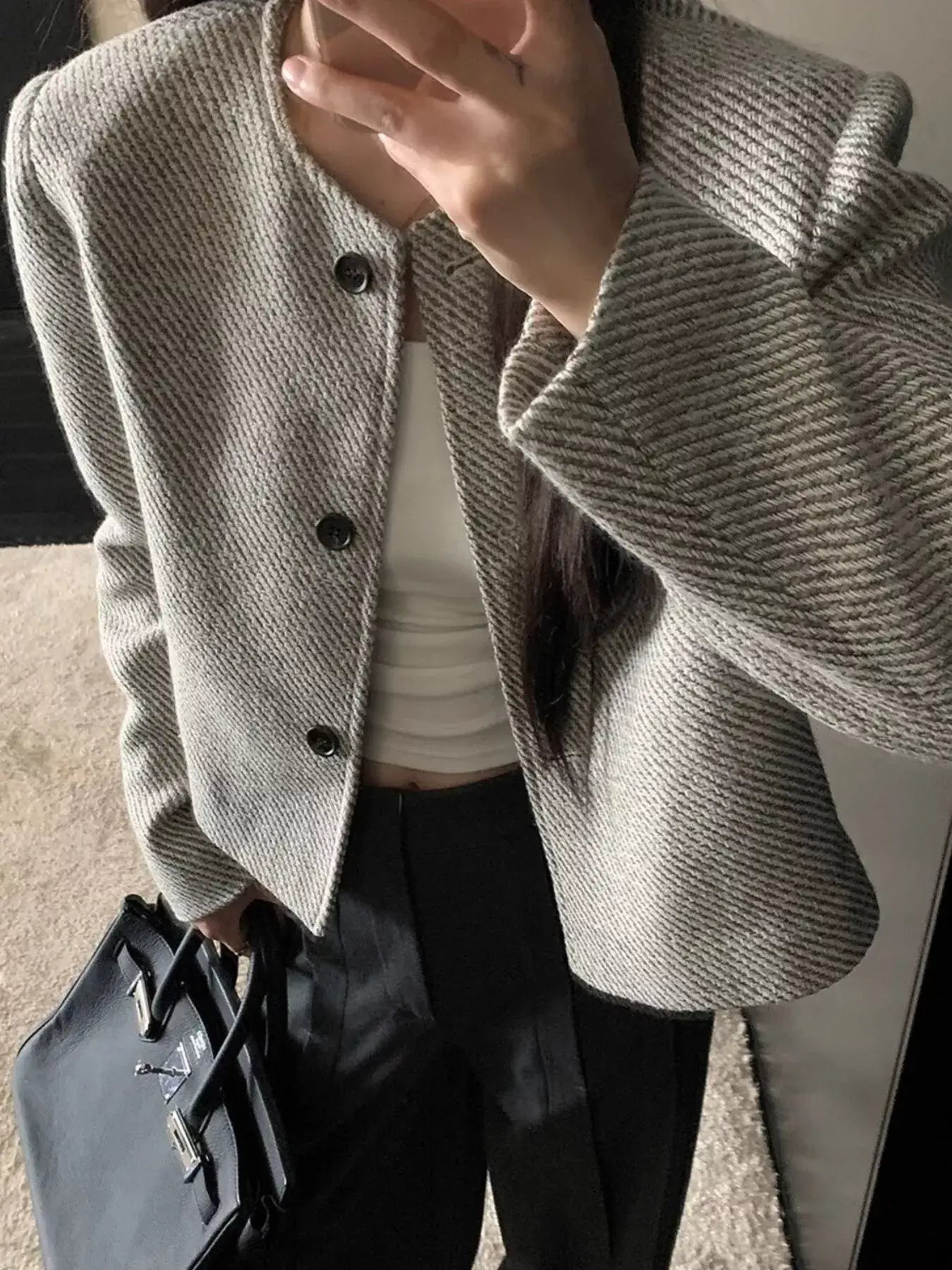 Woman wearing a tweed cropped jacket with buttoned front, holding a bag and wearing a white top and dark pants.