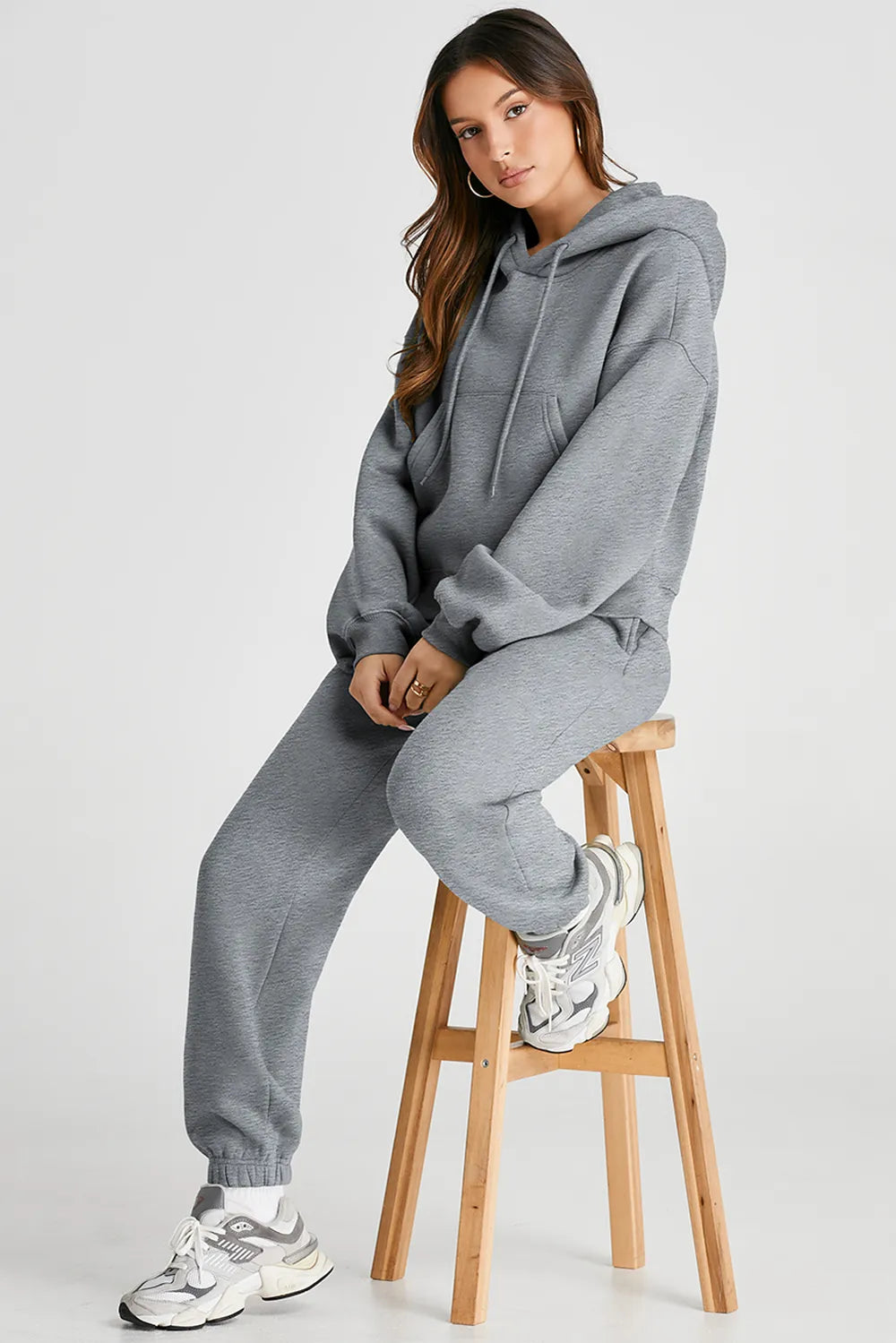 Woman wearing grey Dropped Shoulder Hooded Top and Pants Active Set with drawstring and pockets, sitting on a stool. Casual activewear outfit.