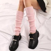 Model wearing Ribbed Knit Leg Warmers in pink, styled with black shoes, sitting on a white sheet for a trendy winter look.