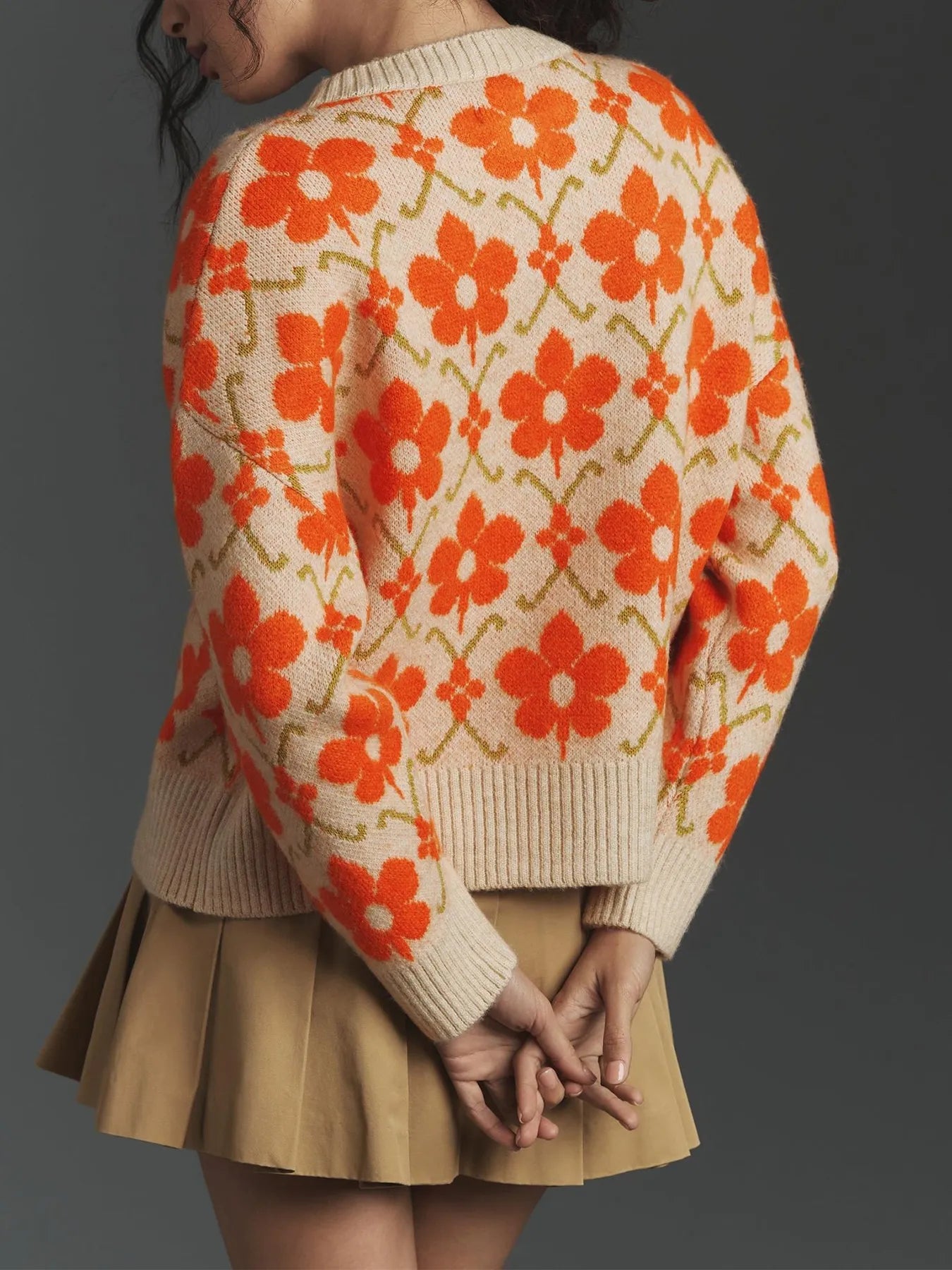Woman wearing floral button-up cardigan with orange flower pattern, long sleeves, and beige background, paired with a khaki skirt.