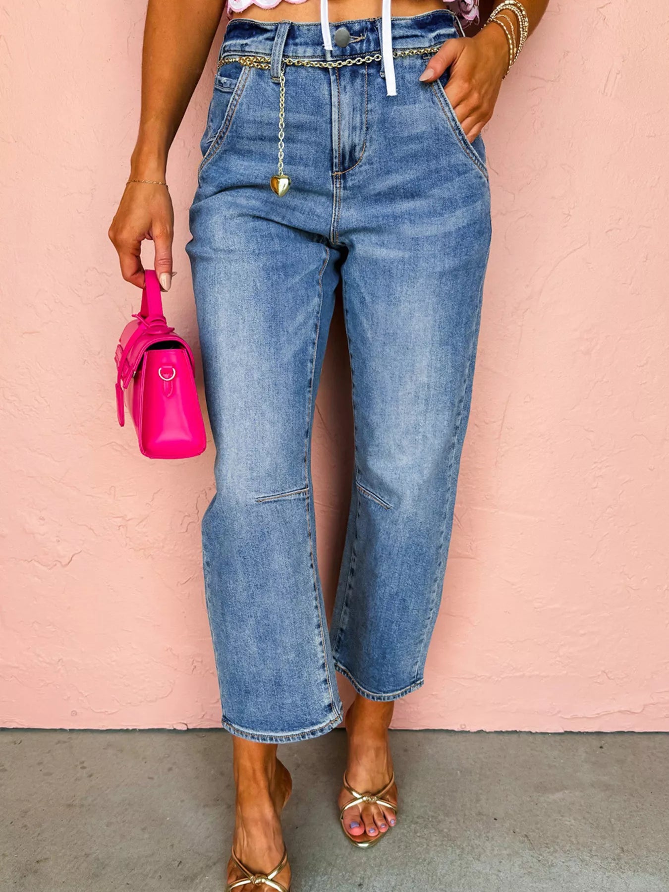 Woman wearing denim seam detail loose fit cropped jeans with a pink bag and sandals against a peach background.