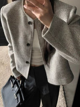 Woman wearing a tweed cropped jacket with buttoned front, holding a bag and wearing a white top and dark pants.