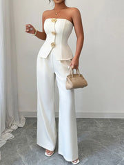 Woman wearing a white tube top and wide-leg pants set, accessorized with a beige handbag and gold buttons, standing in a minimalist setting.