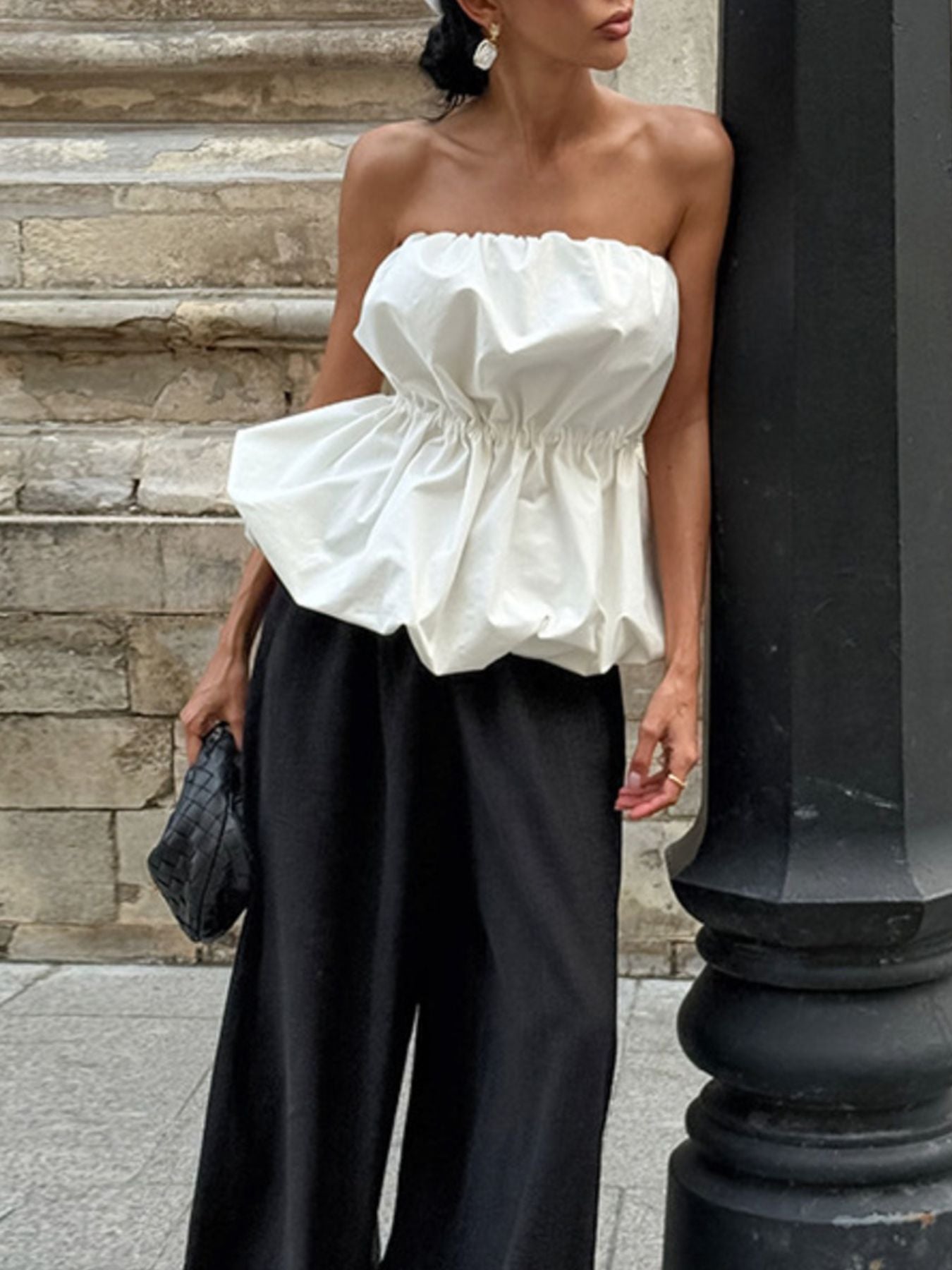 Woman wearing a white Ruched Bubble Hem Tube Top with black trousers, leaning against a stone wall.