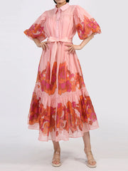Floaty pink button front dress with puff sleeves and floral accents, featuring a belted waist and tiered ruffle hem. Perfect for elegant occasions.
