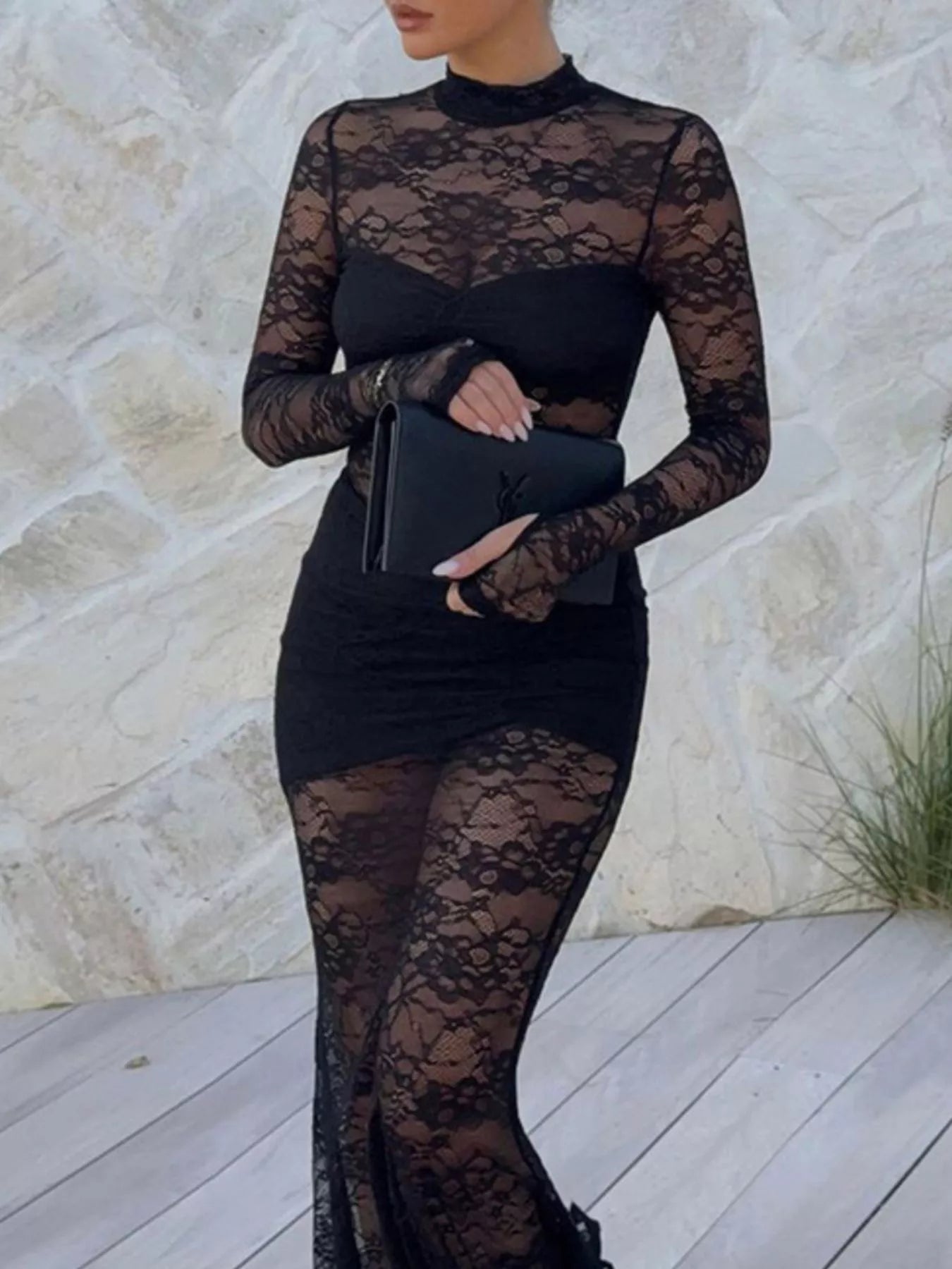 Woman wearing Lace Sheer Mock Neck Long Sleeve Maxi Dress, holding a black clutch against a stone wall background.
